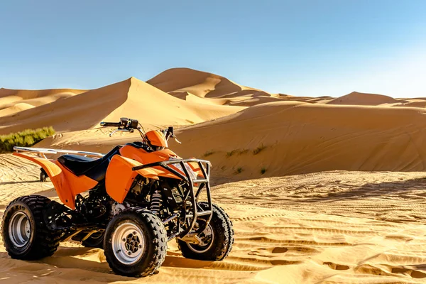 One quad bike orange and black color 4x4 vehicle parked in the Sahara desert, selective focus in foreground, sunny day with golden yellow colored sand dunes, dry bushes and blue sky in blur background