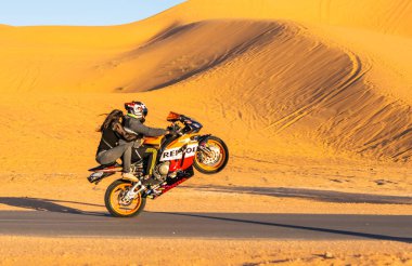Saoura, Cezayir - 28 Aralık 2022 Repsol Honda motosikletini kask takmayan genç bir kızla birlikte Taghit 'te bir çöl yolunda yetiştiren bir motosikletli. Kum tepesi ve mavi gökyüzüyle güneşli bir gün. Hız ve tehlikenin heyecanı.