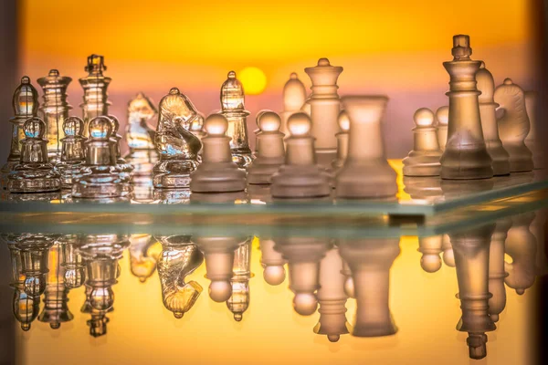 Glass chess pieces reflected with a saturated orange and yellow color solar circle sunset in the blur background. Side view with a horizontal mirror reflexion on the glass table.