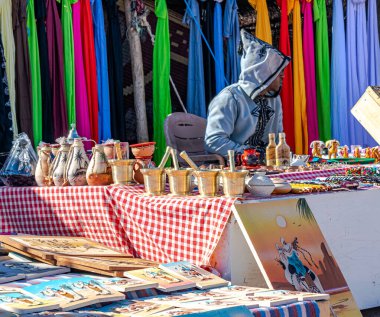Taghit, Cezayir-27 Aralık 2022: Sahra Çölü pazarında hediyelik eşya sergisi. Kapüşonlu yüzü olan tanınmamış bir Tuareg adamı. Renkli eşarplar asıldı, havan topu ve tokmak ve 3D tablolar asıldı