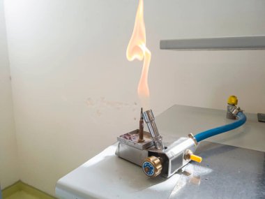 Bir Bunsen ocağı, laboratuvar tezgahında büyük şekilli, turuncu renkli bir alevle tutuştu. Mavi bir gaz tedarik borusu sarı bir gaz kesiciye bağlanıyor. 