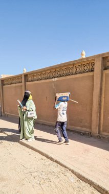 Tamanrasset, Cezayir-10 Ekim 2024: Nane yeşili abaya ve siyah yüz peçesi takmış bir Tuareg kadını ve yüzünü güneşten korumak için kafasına karton bir kutu takmış bir çocuk.