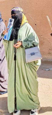 Tamanrasset, Cezayir-10 Ekim 2024: Nane yeşili abaya ve siyah yüz peçesi giymiş bir Tuareg kadını açık havada elinde belgeler, bir akıllı telefon ve parlak güneş ışığı altında açık gri bir el çantası tutuyor.