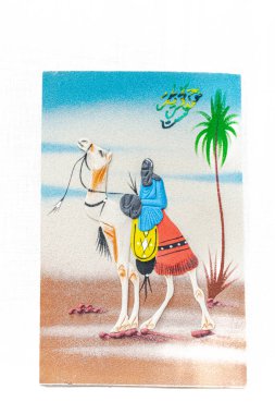 Geleneksel giysiler ve baş örtüsü takan bir Tuareg erkeğinin kumlu bir resmi Sahra Çölü 'nde yürüyen beyaz dromedary deve sürüyor. Arapça bir el yazısı diyor ki 