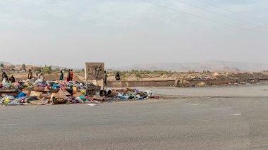 Tamanrasset, Cezayir 22 Mart 2025: Afrikalı erkekler tozlu bir yol kenarında yürürken, el yazısıyla yazılmış bir Arapça ahşap tabelanın yanına taşan çöpler yığılıyor..