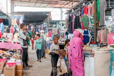 Tamanrasset, Cezayir - 22 Mart 2025: Baharat, tahıl, ayakkabı ve renkli giyim mağazalarını araştıran ailelerle dolu bir pazar sokağı. Kadınlar parlak melhfas giyer ve çocuklar kalabalığın arasında yürürler.