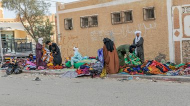 Tamanrasset, Cezayir - 22 Mart 2025: Yürüme alanında gezen satıcılar ve müşterilerin başörtüsü ve djellabas gibi geleneksel kıyafetler giydiği renkli Afrika kumaşları ve giysileri sergilenmektedir.