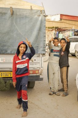 Tamanrasset, Cezayir 22 Mart 2025: Üç gülümseyen çocuk kameraya el işareti ile poz veriyor; biri yalınayak ve ikisi sarılıyor. Tafsit pazarının yakınına bir kamyonet park etmiş.. 