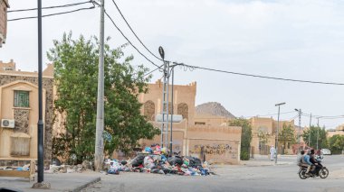 Tamanrasset, Cezayir 22 Mart 2025: Şehirde üç adam motosikletle geçerken, Arapça 'da el yazısıyla yazılmış ahşap bir tabelanın yakınındaki çöp kutusu taşar.. 