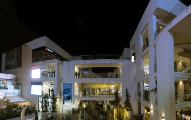 Cezayir - Cezayir - 26 Mart 2025: Geceleri Cheraga 'nın Garden City açık hava alışveriş merkezi yürüyen merdivenler, aydınlatılmış mağaza tabelaları, dekoratif ışıklar, cam balkonlar ve hareket bulanık insanlar.