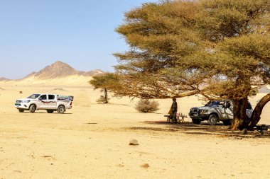 Djanet, Cezayir - 19 Aralık 2021: 4X4 araçlar, tanınmayan tuareg adam, geniş bir çöl bölgesinde Acacia ağacının altında yemek masası. Kuru otlar, sarı renkli kum taşları, kayalık dağ mavisi ve gökyüzü.