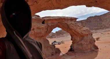 Illizi, Cezayir-21 Aralık 2021: Sahra mağarasında selfie. Doğal kemer, ben, bulutlu gökyüzü, 4x4 arabalar, arka planda sert adamlar. Tanınmayan bir adam kazak takıyor ve yüzü açık değil..