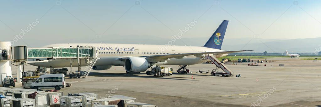 Algiers, Algeria- December 29, 2024: Saudi Arabian airplane Boeing 777 a hallway loading bridge, trucks and vehicles parked on the airport tarmac outdoors, an Air Algrie airplane on the runway afar.