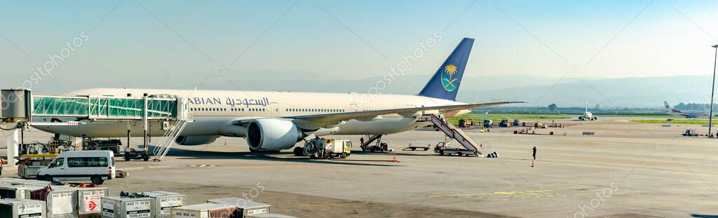 Algiers, Algeria- December 29, 2024: Saudi Arabian airplane Boeing 777 a hallway loading bridge, trucks and vehicles parked on the airport tarmac outdoors, an Air Algrie and another airplane afar.