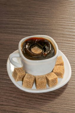 Bir fincan sade kahve ve yüzeye dökülen bir şeker sıçramaya neden oluyor, bir tabak dolusu kahverengi şeker dolu bir fincan tabakta ahşap bir masanın üzerinde servis ediliyor..