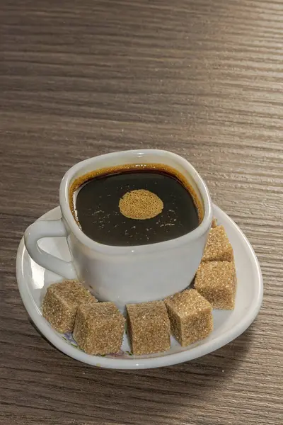 Yüzeyinde hafif köpüklü bir fincan sade kahve, tahtadan bir masanın üzerinde bir sürü kahverengi şeker küpüyle dolu bir fincan beyaz bir fincanda servis edilir..