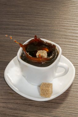 Beyaz bir fincanda servis edilen siyah kahve fincanının dinamik genel manzarası ve tahtadan bir masanın üzerinde kahverengi şekerle servis edilen çay tabağı ve içine küp şekerin düşmesi yüzeyin üzerinde yüksek bir sıçramaya ve damlacıklara neden oluyor.