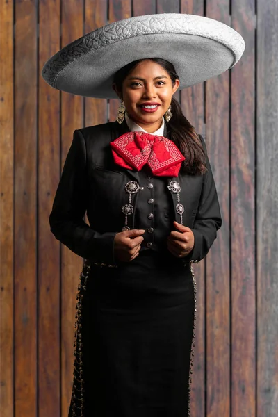 female mexican mariachi woman smiling using a traditional mariachi girl suit on a wooden background. good looking latin hispanic musician feminine mariachi wearing a mexican white hat