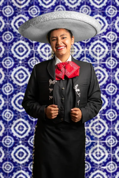 female mexican mariachi woman smiling using a traditional mariachi girl suit on a blue talavera background. good looking latin hispanic musician feminine mariachi wearing a mexican hat
