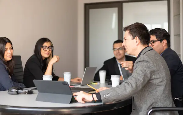 Çalışma alanında çalışan genç beyaz bir adam tabletinin grafiklerini toplantı odasındaki iş arkadaşlarına gösteriyor. Yoğun ofis ortamı, iş adamları...