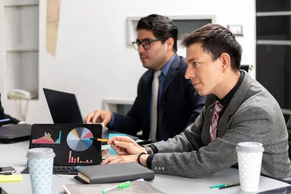 Çalışma alanında çalışan genç beyaz bir adam tabletinin grafiklerini toplantı odasında iş arkadaşına gösteriyor. Ofis ortamları yoğun, iş adamları iş yerinde.