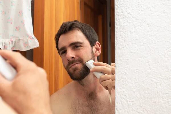 young smiling man grooming shaving and trim the beard with electric razor cutting mustache on the mirror