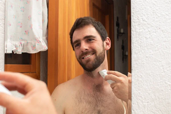 young smiling man grooming shaving and trim the beard with electric razor cutting mustache on the mirror