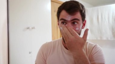 Genç bir adam banyo aynasının önünde yüzüne nemlendirici krem sürüyor. Erkek cilt bakımı, kişisel hijyen, sakallı adam kozmetik maske kullanıyor.
