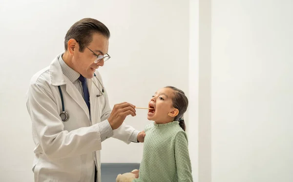 latin doctor checking girl throat using a tongue depressor. Hispanic pediatrician examining kid sore throat, child at doctor\'s office