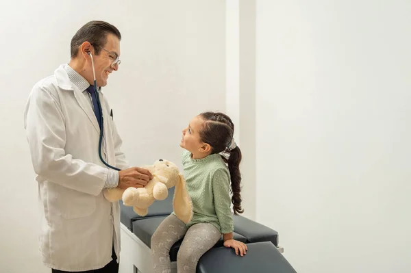 Doktor steteskop kullanarak oyuncak ayının kalp atışlarını dinliyor. Çocuk doktorunun çocuğa güveni tam. Çocuk doktorun ofisinde gülümsüyor. 