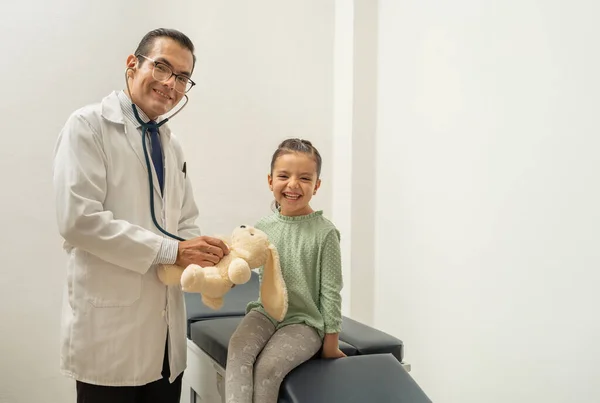 Doktor steteskop kullanarak oyuncak ayının kalp atışlarını dinliyor. Çocuk doktorunun çocuğa güveni tam. Çocuk doktorun ofisinde gülümsüyor. Doktor ve kız kameraya bakıp gülümsüyor. 