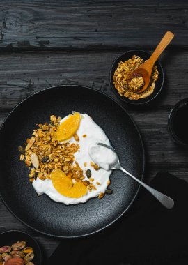 Granola with dried fruits and mandarin slices with Greek yogurt in a black plate on a wooden table.