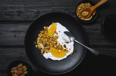 Granola with dried fruits and mandarin slices with Greek yogurt in a black plate on a wooden table.