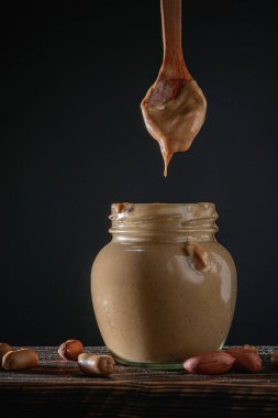 Kalın bir fıstık ezmesi damlası tahta bir kaşıktan cam bir kavanoza düşer. Koyu arkaplan, kaba kompozisyon. Organik, sağlıklı ve minimalist gıda konsepti.