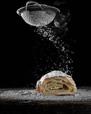 Yukarıdan düşen pudra şekerli taze, pullu kruvasan. Çıtır çıtır tabakalar, siyah arka plan, hareket halindeki yiyecek. Zarif minimalist fırın fotoğrafı.