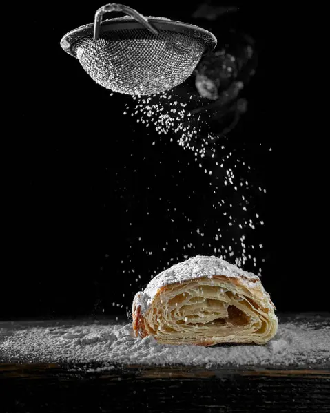 Yukarıdan düşen pudra şekerli taze, pullu kruvasan. Çıtır çıtır tabakalar, siyah arka plan, hareket halindeki yiyecek. Zarif minimalist fırın fotoğrafı.