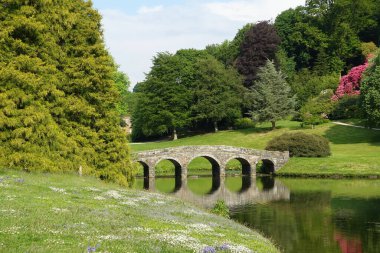 Nehir kıyısında çiçek ve yemyeşil çimlerle dolu bir nehir üzerinde güzel bir taş köprü.