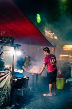 Medium view of Indonesian satay street vendors perform manual grilling over charcoal with heavy white smoke
