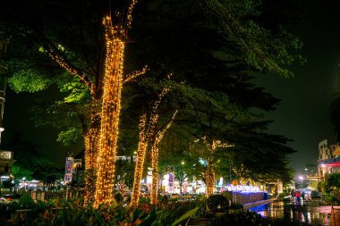 Wide view of decorated medium tree with yellow tumblr lamp in front of mall during the night