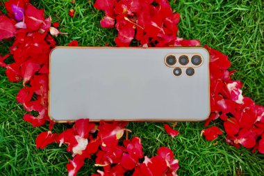 Altın çerçeveli beyaz telefonun arka planı ve güneşli bir günde yeşil çimlerin üzerinde beyaz ve kırmızı çiçek yapraklarıyla birden fazla kamera..