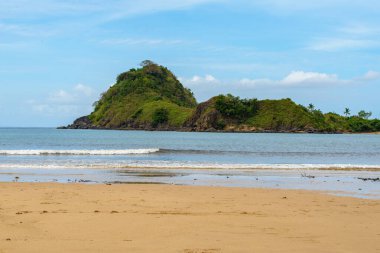 The photo captures a breathtaking view of a serene beach with crystal-clear waters and gentle waves lapping at the shore. The endless stretch of golden sand is dotted with sun umbrellas, beach chairs, and palm trees. The deep blue sea stretches out t