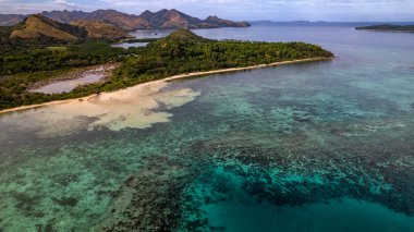 Fotoğraf, berrak suları ve kıyıya vuran hafif dalgalarla sakin bir sahilin nefes kesici görüntüsünü yakalıyor. Sonsuz altın kum taneleri güneş şemsiyeleri, plaj sandalyeleri ve palmiye ağaçları ile kaplıdır.. 