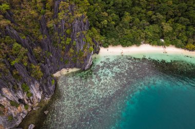 Bu fotoğraf Filipin kıyılarının engebeli güzelliğini yansıtıyor. Ön planda yıpranmış kayalar turkuaz denizden fışkırırken, arka planda el değmemiş beyaz bir kumsal kıyı boyunca kıvrılır..