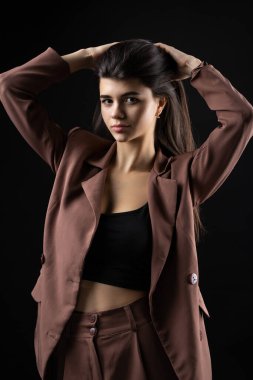 Classic studio portrait of a young brunette dressed in a black top and formal suit, who is sitting on a chair against a black background.