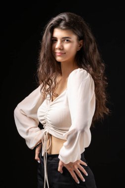 Classic studio portrait of a young brunette in a white loose top, who is sitting on a chair against a black background.