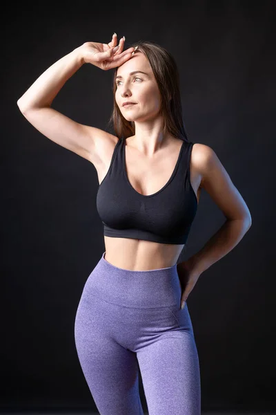 A young healthy girl in a sports top and leggings poses in the studio against a black background.