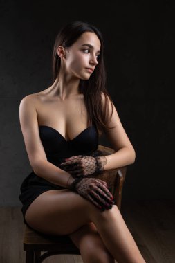 Classic dark studio portrait of a young brunette woman in black clothes who is sitting on a chair.