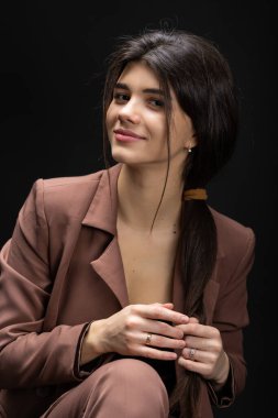 Classic studio portrait of a young brunette dressed in a black top and formal suit, who is sitting on a chair against a black background.