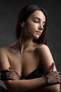 Classic dark studio portrait of a young brunette woman in black clothes who is sitting on a chair.