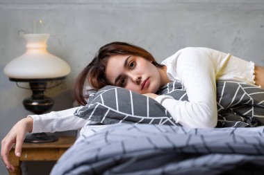 A young attractive girl dressed in white pajamas is resting on a bed in a bedroom in the evening lighting.